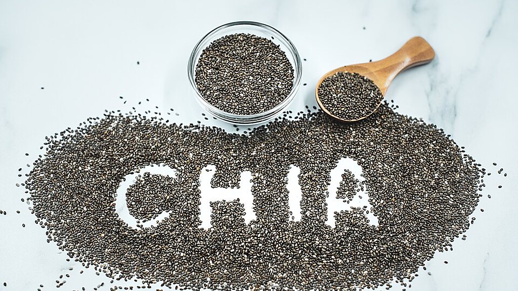 Chia seeds in a glass bowl and on a wooden spoon, with more seeds spelling the word CHIA