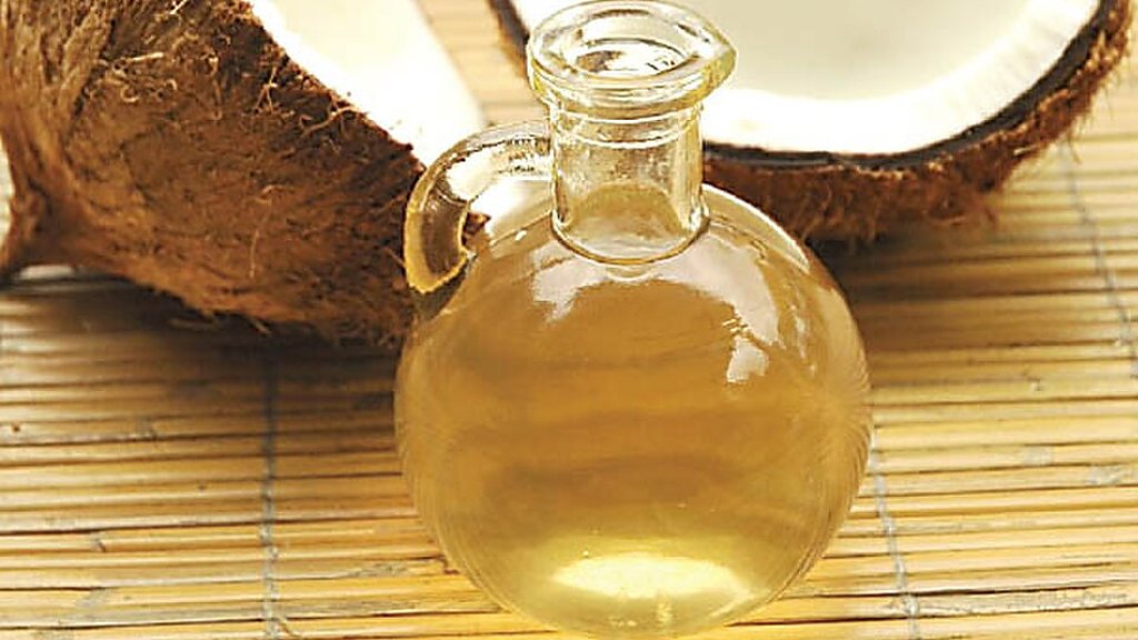 A glass flask of coconut oil in front of two halved coconuts on a bamboo mat